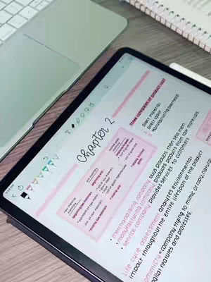 A top-down shot of a digital tablet displaying handwritten notes, placed on a wooden surface. A laptop partially visible in the background adds context to the study environment. The tablet screen is filled with colorful, handwritten notes with pink accents. The composition is clean and organized, suggesting a focus on studying and note-taking.