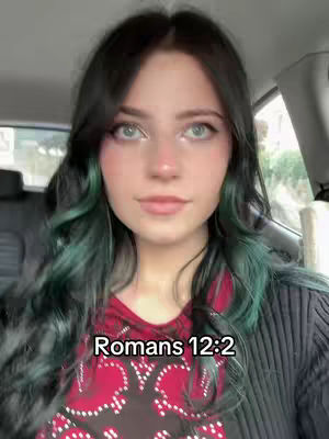 A young woman with long black hair with teal highlights is shown in a selfie taken inside a car. The image is vertically framed. She's wearing a dark jacket over a red patterned shirt, and has evident pink blush on her cheeks. A text overlay at the bottom says 'Romans 12:2'. The image has a soft and well-lit appearance.