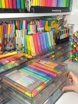 A colorful assortment of pens, markers, and highlighters are neatly organized in clear plastic drawers and stands on a shelf. The image is bright with a variety of colors, creating an aesthetic of a stationery haven.