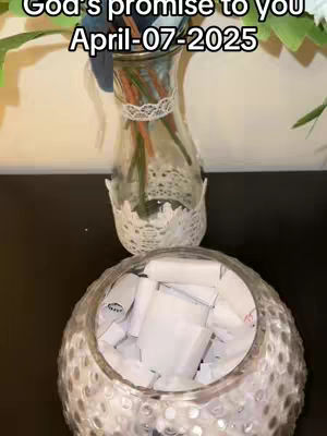 The image shows a clear glass vase filled with water and stems and some leaves, sitting on top of a container. A round, textured glass bowl sits beneath the vase and is filled with numerous white paper scrolls. The background is a dark surface possibly a tabletop.