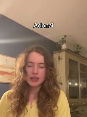 A young woman with long, wavy brown hair is centered in the frame, with her eyes closed and mouth slightly open as if singing. She wears a yellow top. The background includes a wall, the corner of what seems to be a cabinet with some plants on top of it.