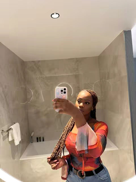 A selfie taken in a bathroom. A young woman with long braided hair holds a phone up to take a picture. She wears a top with various colors, and a black jeans. Behind her is a bathtub, and a mirror reflecting the scene.