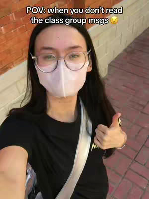 A selfie-style shot of a person wearing glasses, a face mask, and a black shirt, giving a thumbs-up. The background shows a brick wall and an outdoor setting.