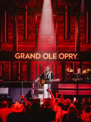 A live performance stage setting with red lighting, a large “GRAND OLE OPRY” sign, and a performer playing a guitar. The stage is lit up with a spotlight, and there's an audience in front.