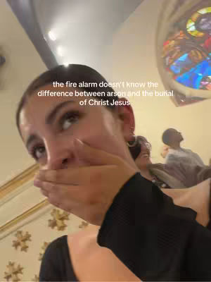 A selfie-style shot of a woman indoors, likely in a church, covering her mouth with her hand. She has a slightly upset expression. The background is blurred, but suggests an ornate interior with architectural details and stained-glass windows. The text overlayed reads a humorous comment about the fire alarm.