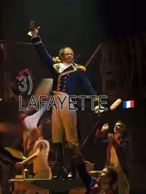 An image of a stage performance. Lafayette, dressed in a costume, is in the center, with one arm raised. His costume is a blue jacket and white pants. Other actors are at the bottom half of the image. There are some text overlays.
