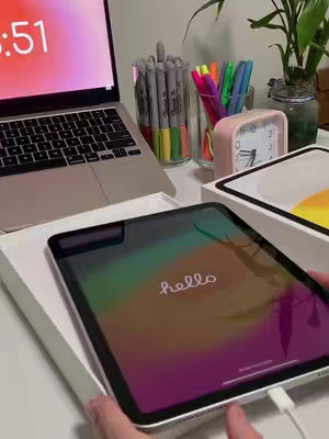 An overhead shot of a workspace with an open laptop, stationary, and an iPad. The iPad displays a colorful background with the word 'hello' on its screen. The scene feels organized and modern with bright accents from the stationary.
