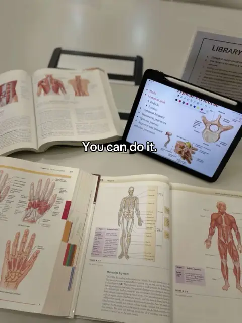 The photo shows a desk with several open anatomy textbooks, an iPad displaying anatomical diagrams with handwritten notes, and a piece of paper with "LIBRARY" printed on it. The composition is from an overhead angle, with the textbooks and iPad arranged to take up the majority of the frame. The background is a plain white surface, possibly a desk. The color palette is dominated by the white background, the cream colored pages of the books, and the black iPad. The overall composition suggests a study environment.
