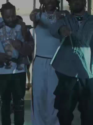 The image shows three men standing in a well-lit indoor space. The man in the center is wearing a light blue tank top and pants and gesturing with his arms. The man to the left is wearing a light blue and white patterned shirt. The man to the right is wearing a blue jacket and black pants. The background consists of simple architecture, suggesting a modern indoor setting.