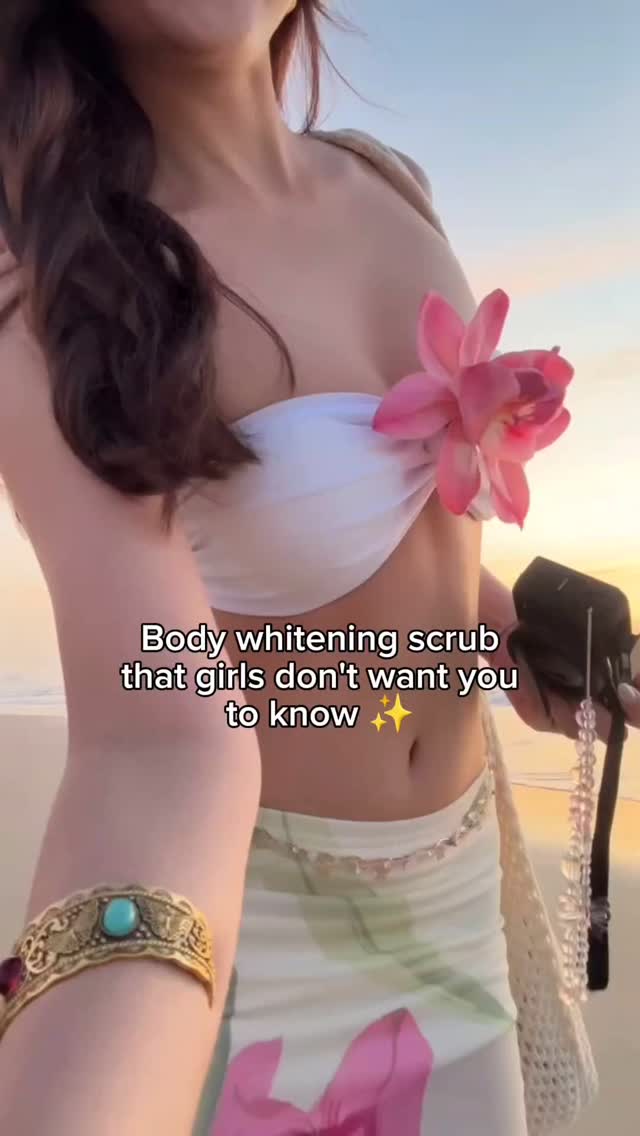 A woman stands on a beach, wearing a white top and a floral skirt. She holds a camera and is adorned with a gold arm cuff and a large pink flower. Text overlayed on the image reads, 'Body whitening scrub that girls don't want you to know'.