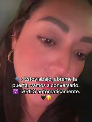 A close-up shot showcases a woman, likely the creator, with her face centered. The upper body is visible, and the background appears blurred, suggesting a close range. She has dark hair and is wearing a small hoop earring. Overlaid text features Spanish phrases related to the Aries zodiac sign and a call to action.