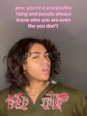 A young woman with curly dark hair is centered in the frame, facing the camera with a neutral expression. She wears an olive green zip-up hoodie with bold, red and white letter designs on the front. The backdrop is blurred, with a soft gradient of pink and light gray colors. The text overlay is pink and positioned above the woman.