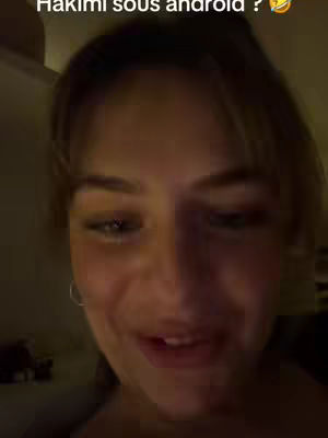 A close-up shot of a young woman's face. She is looking at the camera with a slight smile, and the lighting indicates she is indoors. The background is dark, but there is a hint of a wall or room.