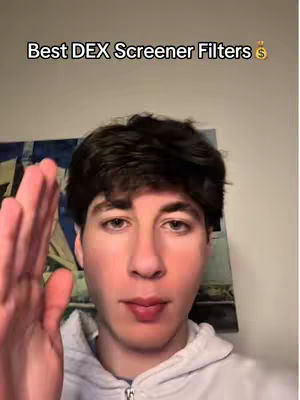 A young man is featured in a selfie-style shot, with his hand raised as if to gesture or pause. The background is a bit blurred, but it seems to have a painting or a large printed image. The lighting feels like it is inside, with a slightly warm tint to the skin tones