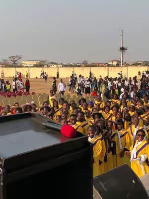 An outdoor gathering with a large crowd of people. The majority is in yellow robes and white or purple scarves. There is a stage or podium in the foreground, partially obscuring a portion of the people. The background shows a clear sky and a fence separating the crowd from a few people walking in the distance.