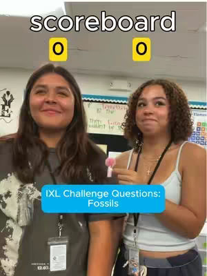 The image is a selfie-style shot featuring two young women in a classroom setting. The main focus is on the two women and a blue rectangle with white font overlay that reads "IXL Challenge Questions: Fossils". Behind them is a whiteboard. The woman on the left has long hair, the woman on the right has curly hair. Above them and in the distance are two yellow squares each with a single number zero.