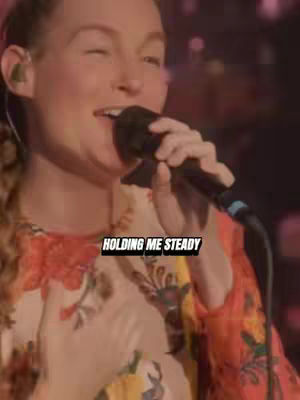 The cover image presents a woman singing with a microphone in hand. She has fair skin and reddish-blonde hair, wearing a colorful patterned top. The background appears to be a blurred performance setting. Text overlayed on the image reads \"HOLDING ME STEADY\" in a bold, white font within a black rectangle.