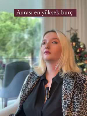 A woman with blonde hair and a leopard-print jacket is looking up. Text overlayed says 'Aurası en yüksek burç'. The background is blurred, featuring a window and a Christmas tree.