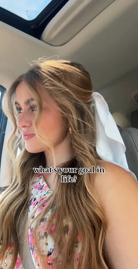 A young woman with long blonde hair is sitting in a car, facing slightly to the side. The scene is lit with natural light, and the background shows a car interior with a sunroof, suggesting an outdoor setting. The woman is wearing a floral patterned top. Text overlay reads, "what's your goal in life?"