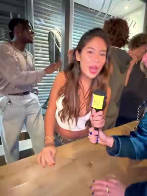 A young woman with long brown hair holding a microphone with a yellow label, speaking with an expressive expression in a crowded outdoor setting. A man is partially visible to her left. Others are in the background.