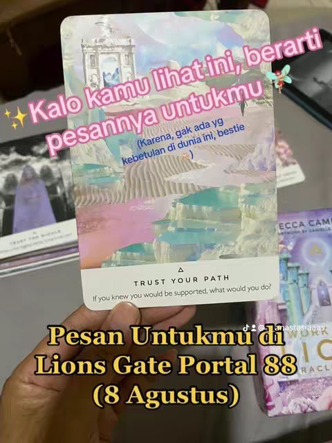 The image shows a hand holding an oracle card in front of other cards and part of a stack of cards with text overlays. The card is predominantly light-colored with an illustration of a building with a blue sky at the top, and a landscape below. Overlayed text uses pink and yellow colors.