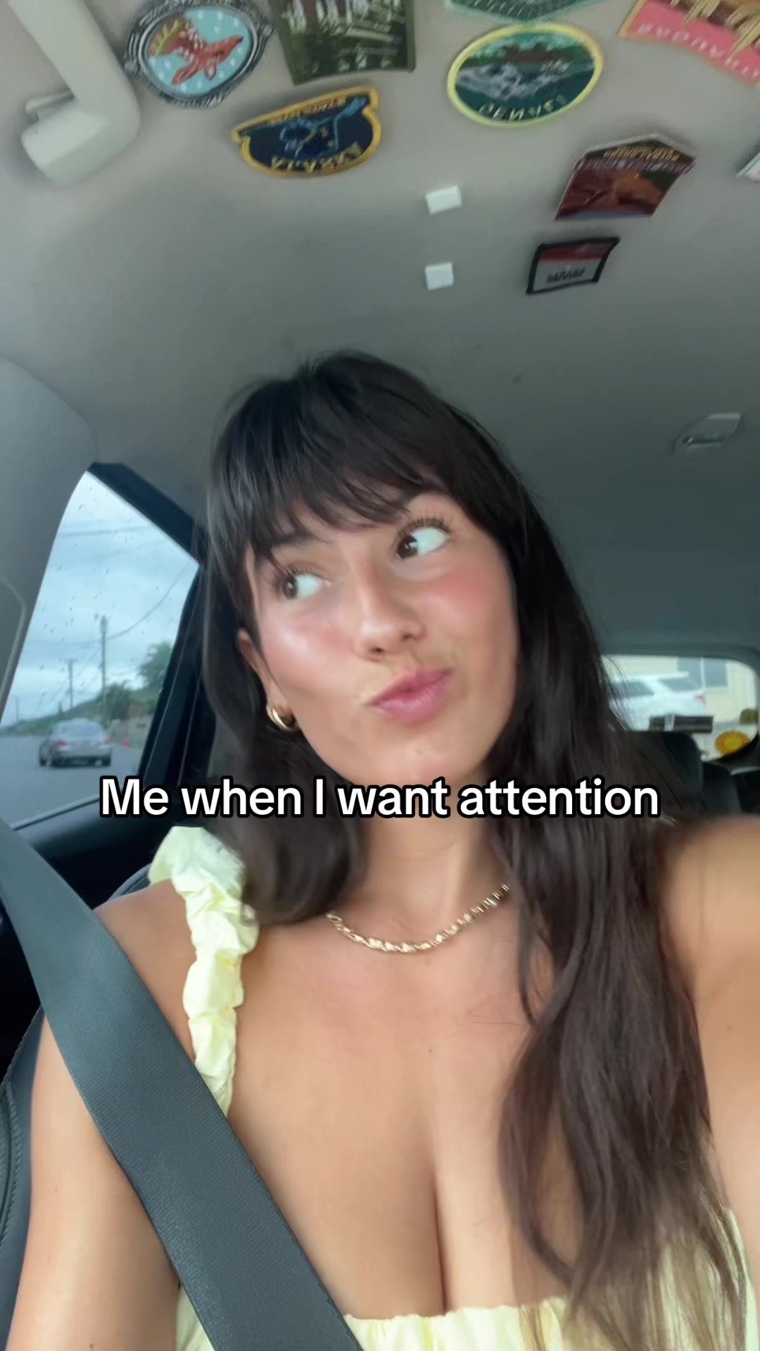A selfie taken from inside a car shows a young woman with brown hair and a yellow top. The woman is the primary focus, with a seatbelt visible across her chest. She has a playful expression, looking to the side with her mouth slightly open. The background shows a road with a car and some stickers on the ceiling.