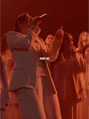 The cover image shows a stage bathed in a warm, red glow. A man wearing a white suit, holding a microphone, is the focal point, singing and gesturing with his right hand. Other people, also in white, are visible in the background, some holding microphones, suggesting a performance. The text overlay says "AND HE".