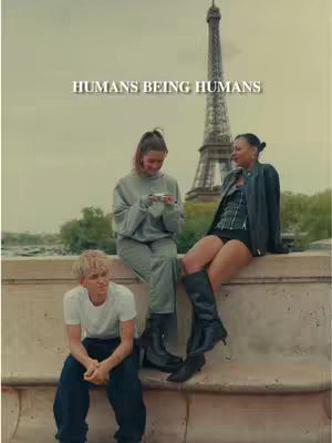 The image features three people sitting on a concrete ledge with the Eiffel Tower in the background. The Eiffel Tower is tall and prominent, while the sky is overcast. The foreground includes three individuals: a young woman in a gray outfit using her phone, another woman in a black jacket and boots with her legs crossed, and a young man in a white t-shirt sitting at the bottom. Text at the top of the image reads 'HUMANS BEING HUMANS'.