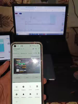 The image shows a person's hand holding a phone displaying a trading app. The phone partially obscures a laptop screen in the background, possibly displaying charts. The setup suggests a cryptocurrency trading environment.