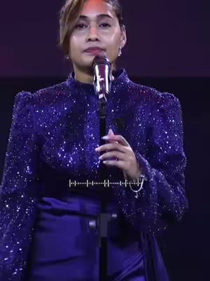 A woman in a sparkling blue jacket stands in front of a microphone, her expression is directed upwards. The background is dark creating a focus on the person. A soundwave graphic is overlaid on the bottom of the image.