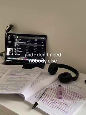 A well-lit, top-down view of a desk setup. The primary focus is a laptop displaying a music streaming app, surrounded by papers, a black headphone, a pen, and a pink highlighter. The background is a simple white wall.