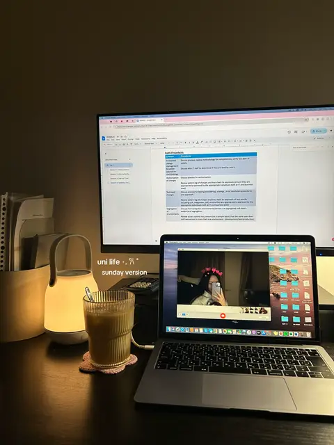 A dimly lit desk setup featuring a laptop displaying a mirrored selfie, a second monitor showing a document, a glowing lamp, and a glass of iced beverage. The scene is a blend of work and leisure, creating a cozy aesthetic.