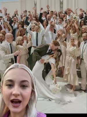 A selfie-style video featuring a woman in the foreground, speaking with an open mouth, with a group of people celebrating a wedding in the background. The background shows a couple kissing with others around them cheering. The overall scene is joyous and celebratory.