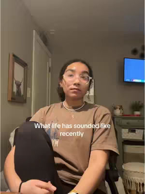 A young woman with glasses sits indoors, angled slightly towards the camera. Her hair is pulled back, and she wears a brown t-shirt. Text overlayed on the image reads, 'What life has sounded like recently.' The background includes a room with artwork and a television.