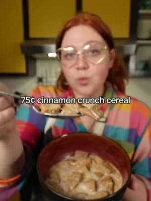 A person with red hair and glasses holds a spoon filled with cereal over a bowl of cereal and milk. The person is wearing a colorful top and the background is a kitchen.