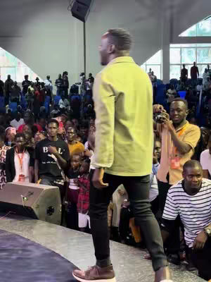 A man in a yellow jacket walks across a stage, facing away from the camera. A large crowd is visible in the background, and a man is taking a photo of the speaker. The lighting makes the faces in the crowd appear dark.