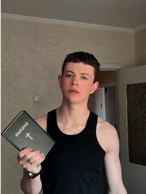 A young man stands indoors, holding a closed book with a cross symbol and the word 'RNINAR' on it. He wears a black tank top and a watch, his gaze fixed on the viewer. The background is a neutral-toned wall with subtle patterns. The lighting appears soft, illuminating his face and upper body.