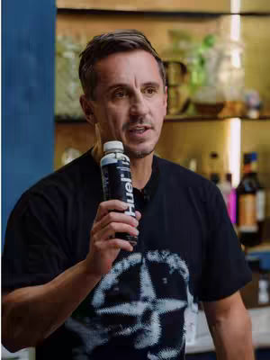 A middle-aged man with short brown hair is holding up a bottle of a dark liquid, appearing to be speaking or presenting the product directly to the camera. He is wearing a black t-shirt with a white emblem on it. The background is blurred, but appears to be a bar or counter setting with bottles and shelves.
