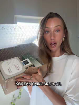 A woman with long blonde hair is holding a box of cosmetic products, appearing surprised. The scene is indoors, with a window and a natural lighting setting. The image has text overlaying the bottom, reading 'KOREAN SKINCARE HAUL'.