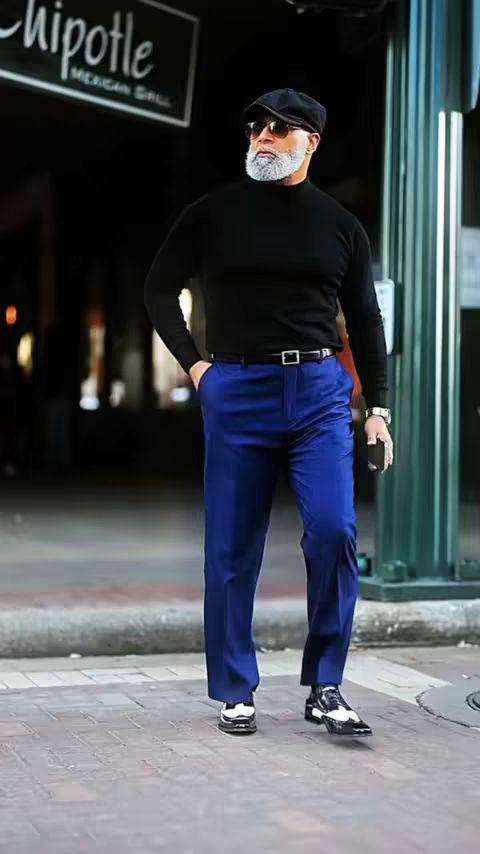 A well-dressed, older man stands on a brick sidewalk with his hand in his pocket. He wears a black turtleneck, blue pants, and two-tone shoes. A Chipotle sign is visible in the background.