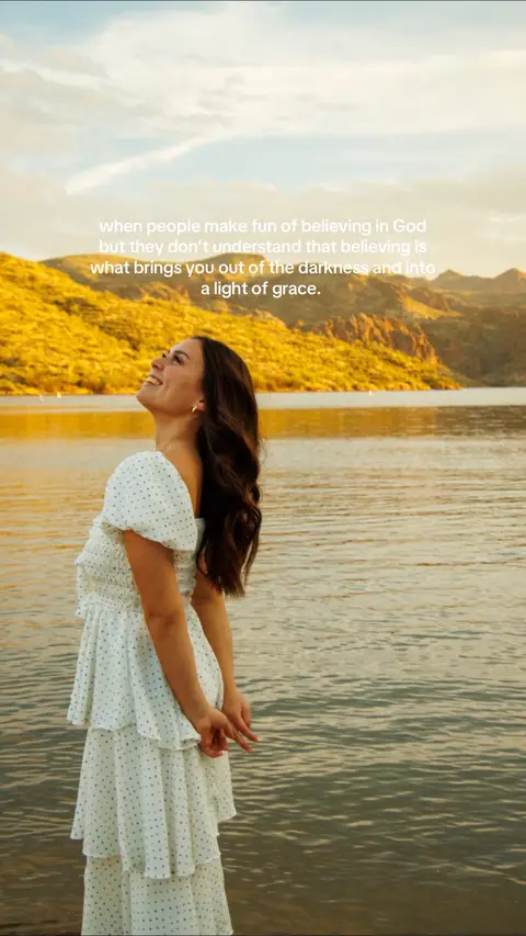 A woman with long brown hair stands near a lake, looking upwards and smiling with a backdrop of golden mountains and a bright sky. She wears a white dress with blue polka dots.