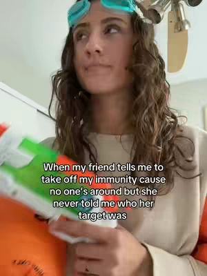A woman with curly brown hair is looking upwards and to the left. She is wearing a light-colored long-sleeved shirt and blue swimming goggles on her forehead. She is holding a water gun. The background is a light-colored room with a partial view of a ceiling fan and a wall. Text is overlaid on the image.