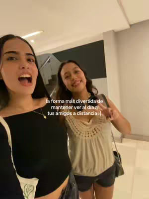 Two young women are captured in a selfie-style shot, smiling and interacting within what appears to be an indoor space. The woman on the left has dark hair, and is opening her mouth to talk excitedly. The woman on the right is smiling and gesturing with her fingers. Overlaid text suggests the video's purpose is to promote an app for staying in touch with friends.