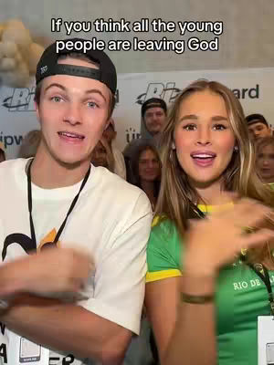 A cover image featuring a young man and woman smiling and speaking animatedly. The man wears a black cap, a white t-shirt, and a black lanyard. The woman wears a green and yellow top. Behind them, other people are visible, and there is text overlayed at the top.