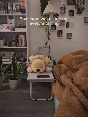 A room scene with a bed covered in a brown blanket, a small table with a Winnie-the-Pooh plushie resting on it, and a bookshelf. The text overlay reads 'Pov: mom wakes me up every morning'.