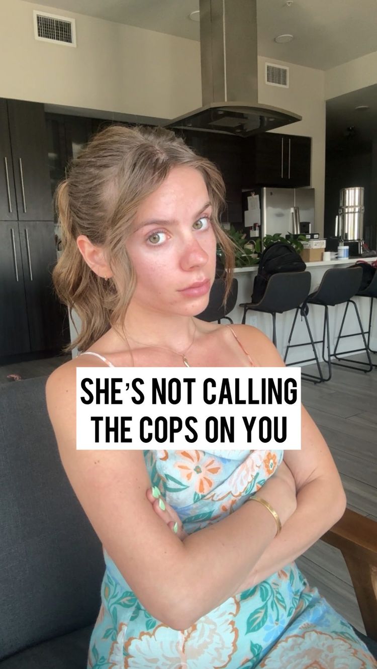 A woman with light brown hair and fair skin sits in a chair, looking directly at the camera with a neutral expression. The background shows a modern kitchen with dark cabinetry, and a text overlay reads, "SHE'S NOT CALLING THE COPS ON YOU." She wears a light-colored top and has her arms crossed.