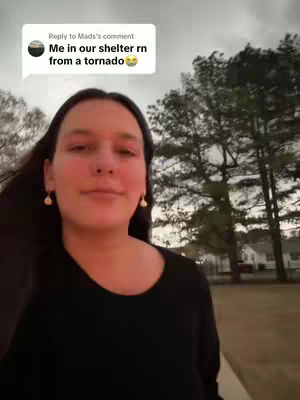 A woman is standing outside, smiling, with a residential area and trees in the background under an overcast sky. A text overlay indicates she is in a shelter from a tornado.