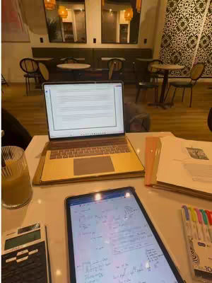 A well-lit interior shot of a cafe or study area, where the focus is on a workspace set up which consists of a laptop and a tablet with study materials laid out on a white table. Various pens, a calculator, and a glass of drink are spread around. The background shows a blurry cafe setting.