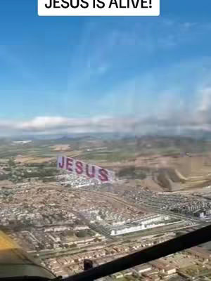 An aerial view from a cockpit, looking out over a city and landscape. A banner with the word 'JESUS' is being towed by an airplane. The sky is blue with some clouds.