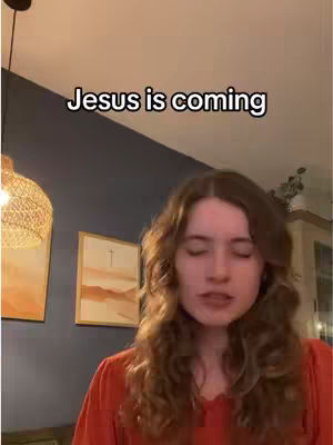A woman with long brown hair, wearing a red top, sings with her eyes closed in front of a dark blue wall. Above her head the words "Jesus is coming" appear in a white rectangular banner. In the background a lamp, artwork, and some greenery add context to the scene.